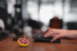 A colorful spinning top with intricate designs on its surface is in focus, sitting on a wooden table. In the background, a hand is typing on a keyboard, blurred out to emphasize the top.