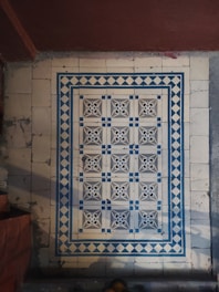 Intricate floor tiles with a decorative pattern, featuring symmetrical design elements in blue and white. The tiles are framed by a diamond border, creating a visually striking appearance. The floor appears aged and shows signs of wear.