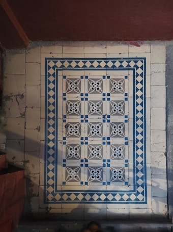 Intricate floor tiles with a decorative pattern, featuring symmetrical design elements in blue and white. The tiles are framed by a diamond border, creating a visually striking appearance. The floor appears aged and shows signs of wear.