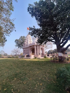 A traditional temple with intricate details stands surrounded by a lush green lawn and trees. The temple has a flag atop, signaling its cultural significance. There are steps leading up to the entrance, and the scene is calm with fallen leaves scattered across the grass.