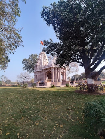 A traditional temple with intricate details stands surrounded by a lush green lawn and trees. The temple has a flag atop, signaling its cultural significance. There are steps leading up to the entrance, and the scene is calm with fallen leaves scattered across the grass.