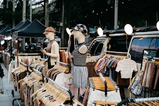 A street market stall displays various clothes on hangers. Two mannequins, dressed in casual outfits, showcase garments near a van. String lights hang above, and there are several black tents set up further along the street.