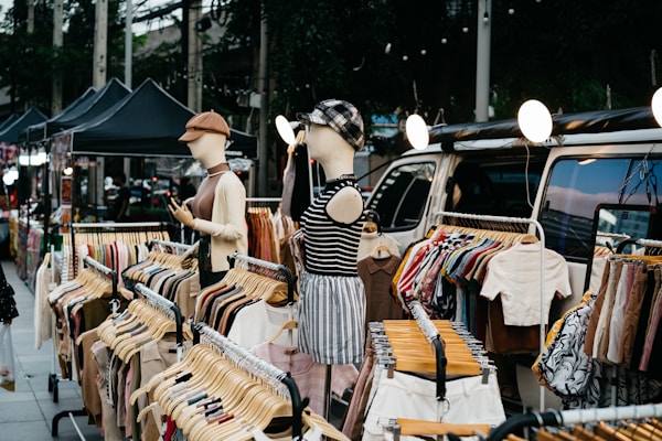 A street market stall displays various clothes on hangers. Two mannequins, dressed in casual outfits, showcase garments near a van. String lights hang above, and there are several black tents set up further along the street.
