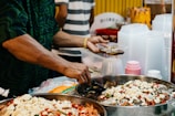 A person is preparing and serving food from large metal bowls filled with various ingredients, including chopped white pieces and colorful chili peppers. The person wears a patterned dark shirt and a gold bracelet, and is using a small plastic container. Nearby are stacks of plastic cups and containers, indicating a food stall or market setting.