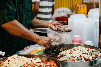 A person is preparing and serving food from large metal bowls filled with various ingredients, including chopped white pieces and colorful chili peppers. The person wears a patterned dark shirt and a gold bracelet, and is using a small plastic container. Nearby are stacks of plastic cups and containers, indicating a food stall or market setting.