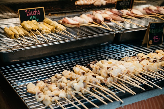 Various types of skewered foods are placed on a metal grill. The skewers are filled with what appears to be different meats and seafood. The background shows signs with prices written in a foreign language.