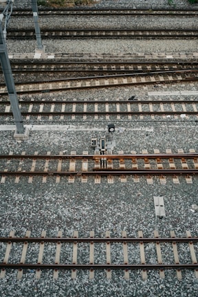 An aerial view of multiple parallel railway tracks laid on a bed of gravel. The tracks are evenly spaced and supported by wooden sleepers. A metal pole is visible to the left side of the image, possibly part of the railway infrastructure.