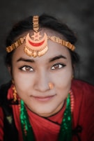 A person wearing traditional jewelry with a prominent chain headpiece featuring crescent and star designs. The attire includes vibrant colors and jewelry, such as a nose ring and necklace. The background is softly blurred, drawing focus to the individual's detailed facial features and adornments.