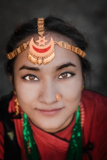 A person wearing traditional jewelry with a prominent chain headpiece featuring crescent and star designs. The attire includes vibrant colors and jewelry, such as a nose ring and necklace. The background is softly blurred, drawing focus to the individual's detailed facial features and adornments.