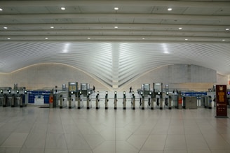 A modern transportation hub with sleek, curved architectural elements. Multiple electronic turnstiles are arranged in a line, with high ceilings and ample lighting enhancing the spaciousness. There are signs for ticketing and a banner indicating an event, with a few people visible in the distance.