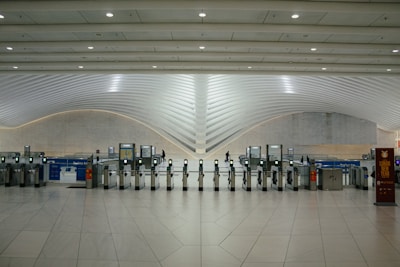 A modern transportation hub with sleek, curved architectural elements. Multiple electronic turnstiles are arranged in a line, with high ceilings and ample lighting enhancing the spaciousness. There are signs for ticketing and a banner indicating an event, with a few people visible in the distance.