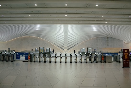A modern transportation hub with sleek, curved architectural elements. Multiple electronic turnstiles are arranged in a line, with high ceilings and ample lighting enhancing the spaciousness. There are signs for ticketing and a banner indicating an event, with a few people visible in the distance.