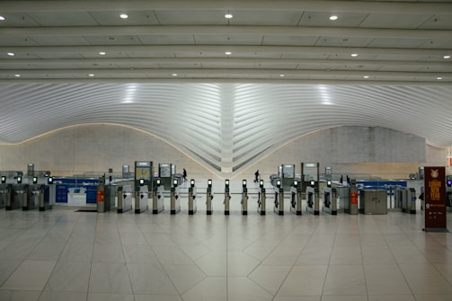 A modern transportation hub with sleek, curved architectural elements. Multiple electronic turnstiles are arranged in a line, with high ceilings and ample lighting enhancing the spaciousness. There are signs for ticketing and a banner indicating an event, with a few people visible in the distance.
