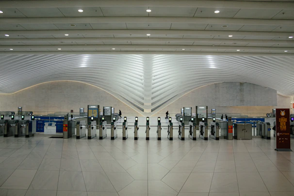 A modern transportation hub with sleek, curved architectural elements. Multiple electronic turnstiles are arranged in a line, with high ceilings and ample lighting enhancing the spaciousness. There are signs for ticketing and a banner indicating an event, with a few people visible in the distance.