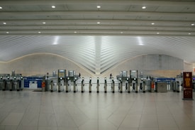 A modern transportation hub with sleek, curved architectural elements. Multiple electronic turnstiles are arranged in a line, with high ceilings and ample lighting enhancing the spaciousness. There are signs for ticketing and a banner indicating an event, with a few people visible in the distance.