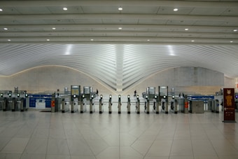 A modern transportation hub with sleek, curved architectural elements. Multiple electronic turnstiles are arranged in a line, with high ceilings and ample lighting enhancing the spaciousness. There are signs for ticketing and a banner indicating an event, with a few people visible in the distance.