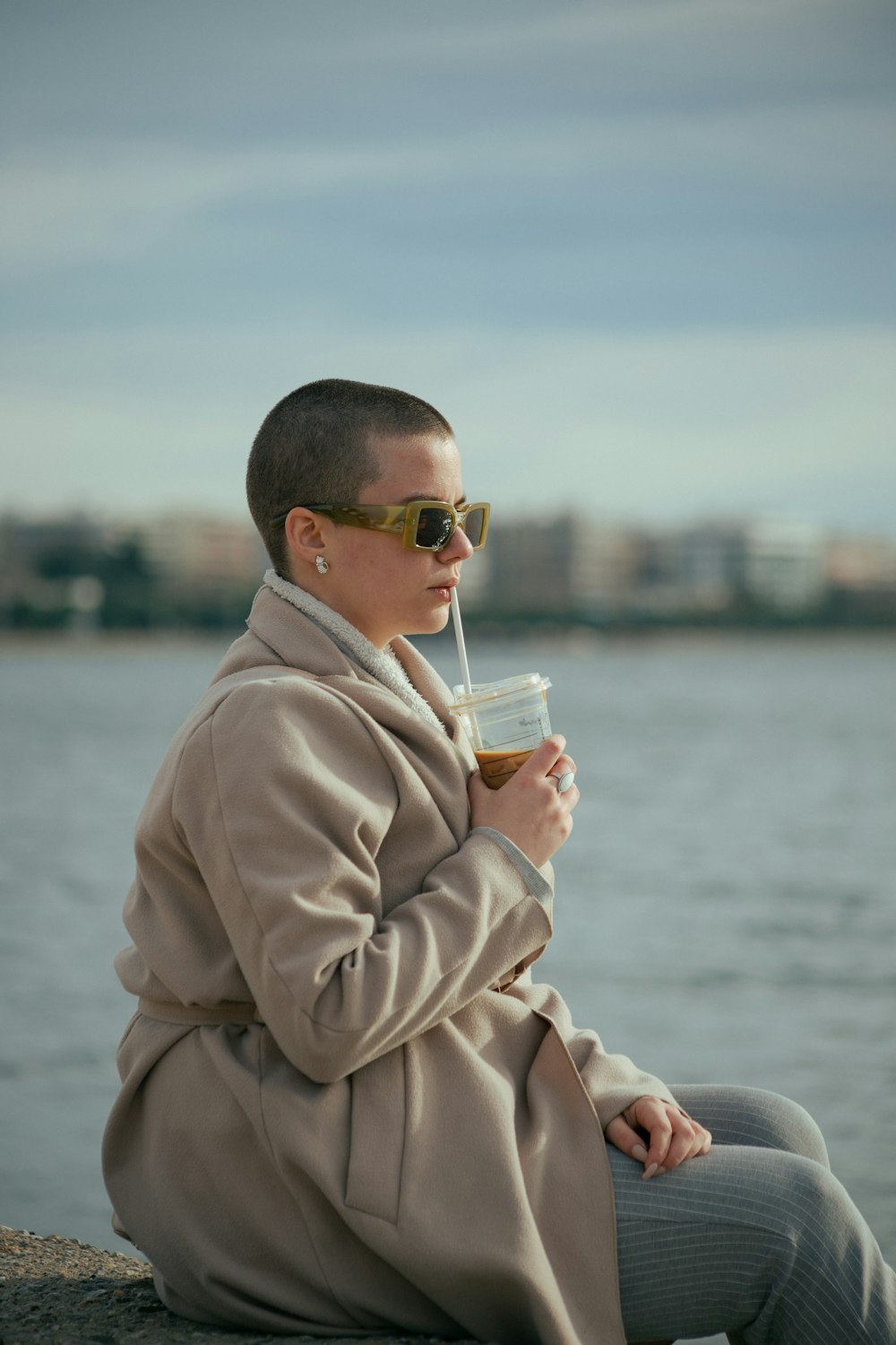 a woman in a trench coat and sunglasses sitting by the water