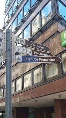 A metal post with directional signs pointing towards Váci Street, Pest Redoute, and Danube Promenade, set against the background of a modern glass building. Reflections of nearby structures are visible in the windows, and there are clear skies reflected above.