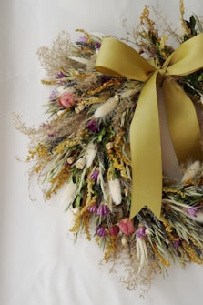 A decorative wreath composed of a variety of dried flowers and plants, featuring a prominent large yellow bow. The arrangement includes pops of pink, purple, white, and green, with textures from grasses and foliage adding depth.
