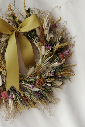 A decorative wreath made of dried flowers and grasses, adorned with a large golden ribbon tied into a bow. The wreath features various elements such as purple thistles, seed pods, and green foliage intertwined with yellow dried blooms. The backdrop is a soft white fabric, providing contrast to the natural tones of the wreath.