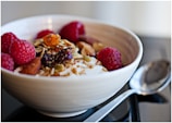 A close-up of a bowl filled with creamy yogurt topped with raspberries, nuts, seeds, and dried fruit. A shiny metal spoon is placed beside the bowl on a smooth surface.