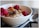 A close-up of a bowl filled with creamy yogurt topped with raspberries, nuts, seeds, and dried fruit. A shiny metal spoon is placed beside the bowl on a smooth surface.