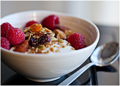 A close-up of a bowl filled with creamy yogurt topped with raspberries, nuts, seeds, and dried fruit. A shiny metal spoon is placed beside the bowl on a smooth surface.