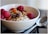 A close-up of a bowl filled with creamy yogurt topped with raspberries, nuts, seeds, and dried fruit. A shiny metal spoon is placed beside the bowl on a smooth surface.