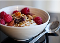 A close-up of a bowl filled with creamy yogurt topped with raspberries, nuts, seeds, and dried fruit. A shiny metal spoon is placed beside the bowl on a smooth surface.