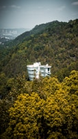 A modern, white, glass-structured building is nestled among dense, lush green forests on a hillside. The landscape is dominated by varying shades of green with a cityscape faintly visible in the distant background.