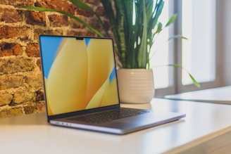 A sleek laptop with a vibrant screen sits open on a white desk in a modern workspace. Behind it, a tall green plant in a white pot adds a natural touch, and a textured brick wall provides a rustic background. Natural light filters in from large windows.