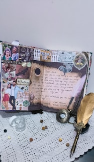 A creatively decorated scrapbook page featuring photos, handwritten notes, and artistic embellishments. The page includes a prominent love letter, images of a couple, and the word 'Erkenci Kus'. The surrounding elements include a feather pen, a small glass jar, coins, and an ornate pocket watch lying on a lace tablecloth.