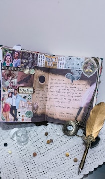 A creatively decorated scrapbook page featuring photos, handwritten notes, and artistic embellishments. The page includes a prominent love letter, images of a couple, and the word 'Erkenci Kus'. The surrounding elements include a feather pen, a small glass jar, coins, and an ornate pocket watch lying on a lace tablecloth.