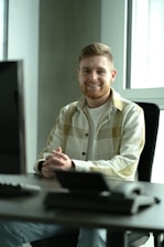 A bearded person with short hair, wearing a plaid shirt over a white t-shirt, is seated at a desk in an office setting. The person is smiling and has their hands clasped in front of them on the desk. There is a computer monitor in the foreground on the left side of the image and large windows in the background allowing natural light to come in.