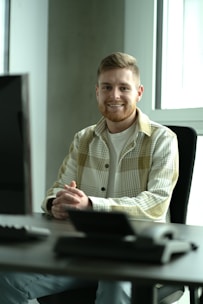 A bearded person with short hair, wearing a plaid shirt over a white t-shirt, is seated at a desk in an office setting. The person is smiling and has their hands clasped in front of them on the desk. There is a computer monitor in the foreground on the left side of the image and large windows in the background allowing natural light to come in.