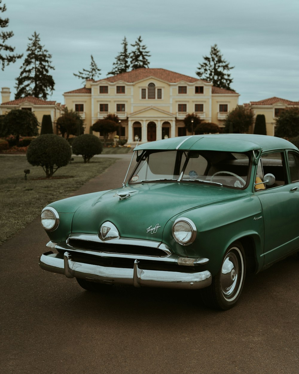 an old green car parked in front of a large house