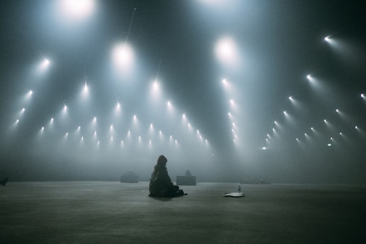 A large, dimly lit space with numerous overhead lights emitting soft glows, creating a surreal atmosphere. In the middle distance, a solitary figure sits on the floor, surrounded by a vast emptiness. There is a strong sense of stillness and solitude.