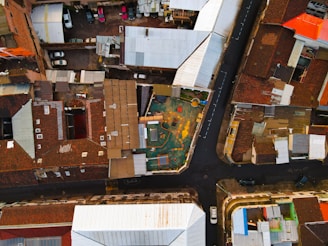 An aerial view of an urban area featuring a central playground surrounded by various residential buildings with tiled roofs. The roads form distinct boundaries and reveal parked cars along the streets and inside driveways. The playground area appears colorful and well-defined in the midst of densely packed structures.