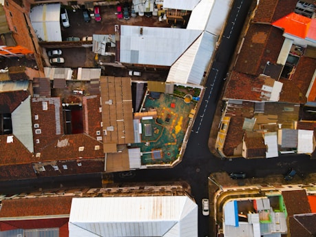 An aerial view of an urban area featuring a central playground surrounded by various residential buildings with tiled roofs. The roads form distinct boundaries and reveal parked cars along the streets and inside driveways. The playground area appears colorful and well-defined in the midst of densely packed structures.