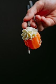 A hand holding a piece of smoked salmon adorned with a generous swirl of cream cheese or similar topping, using chopsticks against a dark background.