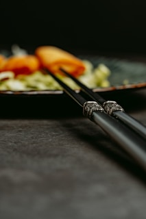 A pair of ornate chopsticks rests on a dark surface, with a blurred background showing slices of orange-colored food, possibly salmon, and some green vegetables on a plate.