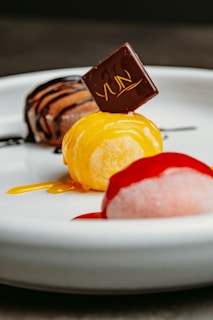 A dessert plate features three round mochi ice creams, each topped with a different sauce. The front mochi is pink with a red sauce, the middle is yellow with a chocolate garnish, and the back is brown with chocolate drizzle.