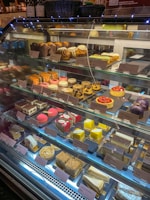 A display case filled with a variety of colorful pastries and cakes, neatly arranged on shelves. The assortment includes cakes with various toppings, macarons, eclairs, and other desserts, each labeled with a price tag.