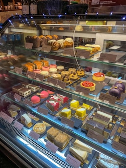 A display case filled with a variety of colorful pastries and cakes, neatly arranged on shelves. The assortment includes cakes with various toppings, macarons, eclairs, and other desserts, each labeled with a price tag.