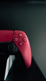 A close-up view of a red gaming controller resting on a dark surface. The focus is on the buttons, including the familiar shapes of a square, triangle, circle, and cross.