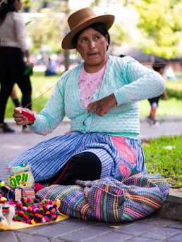 A woman is sitting on a paved walkway, focused on her craftwork. She is dressed in a traditional outfit with vibrant colors, wearing a hat adorned with a feather. Her attention is on the yarn she is working with. Next to her are colorful textiles and small handmade items displayed for sale, and a bag of snacks is visible. The background is a blurred park setting with greenery.