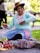 A woman is sitting on a paved walkway, focused on her craftwork. She is dressed in a traditional outfit with vibrant colors, wearing a hat adorned with a feather. Her attention is on the yarn she is working with. Next to her are colorful textiles and small handmade items displayed for sale, and a bag of snacks is visible. The background is a blurred park setting with greenery.