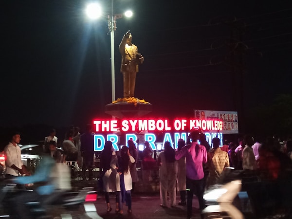 A statue atop a pedestal is illuminated by bright lights. In front of the statue, there is a large neon sign displaying the words 'THE SYMBOL OF KNOWLEDGE DR B R AMBEDKAR'. A crowd of people is gathered around the area, some taking photographs. The scene is set in a public space at night.