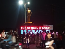 A statue atop a pedestal is illuminated by bright lights. In front of the statue, there is a large neon sign displaying the words 'THE SYMBOL OF KNOWLEDGE DR B R AMBEDKAR'. A crowd of people is gathered around the area, some taking photographs. The scene is set in a public space at night.