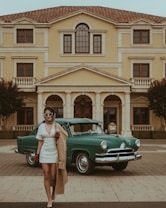 A woman with sunglasses, a headscarf, and a short white dress stands in front of a vintage green car, posing stylishly. The background features a grand, beige-colored building with a classical architectural style, complete with arched windows and pillars. The scene exudes an air of elegance and nostalgia.